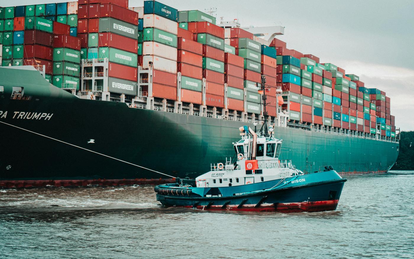 a tug boat pulling a large container ship by Mika Baumeister courtesy of Unsplash.