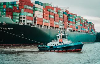 a tug boat pulling a large container ship by Mika Baumeister courtesy of Unsplash.